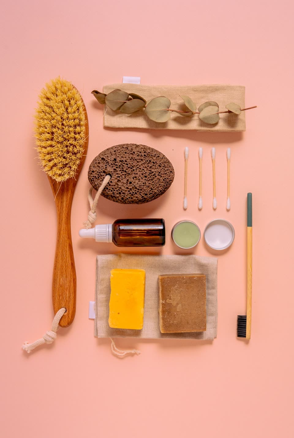 A wooden body brush, pumice, bamboo cotton buds and bar soaps laid out on pink