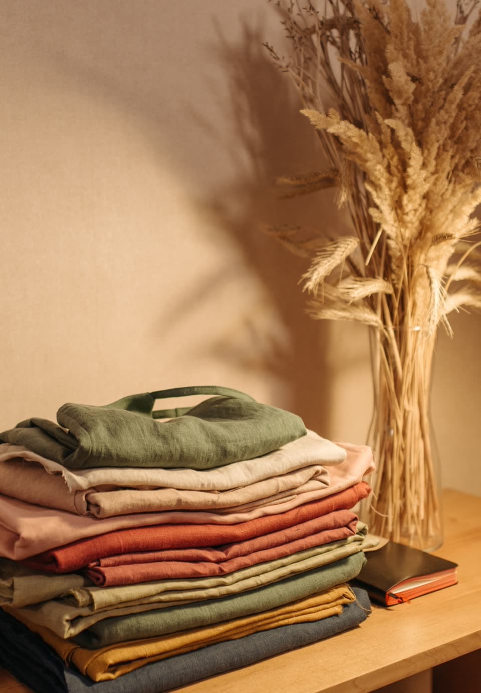 A stack of folded clothes in green, rust and cream beside a vase of dried wheat